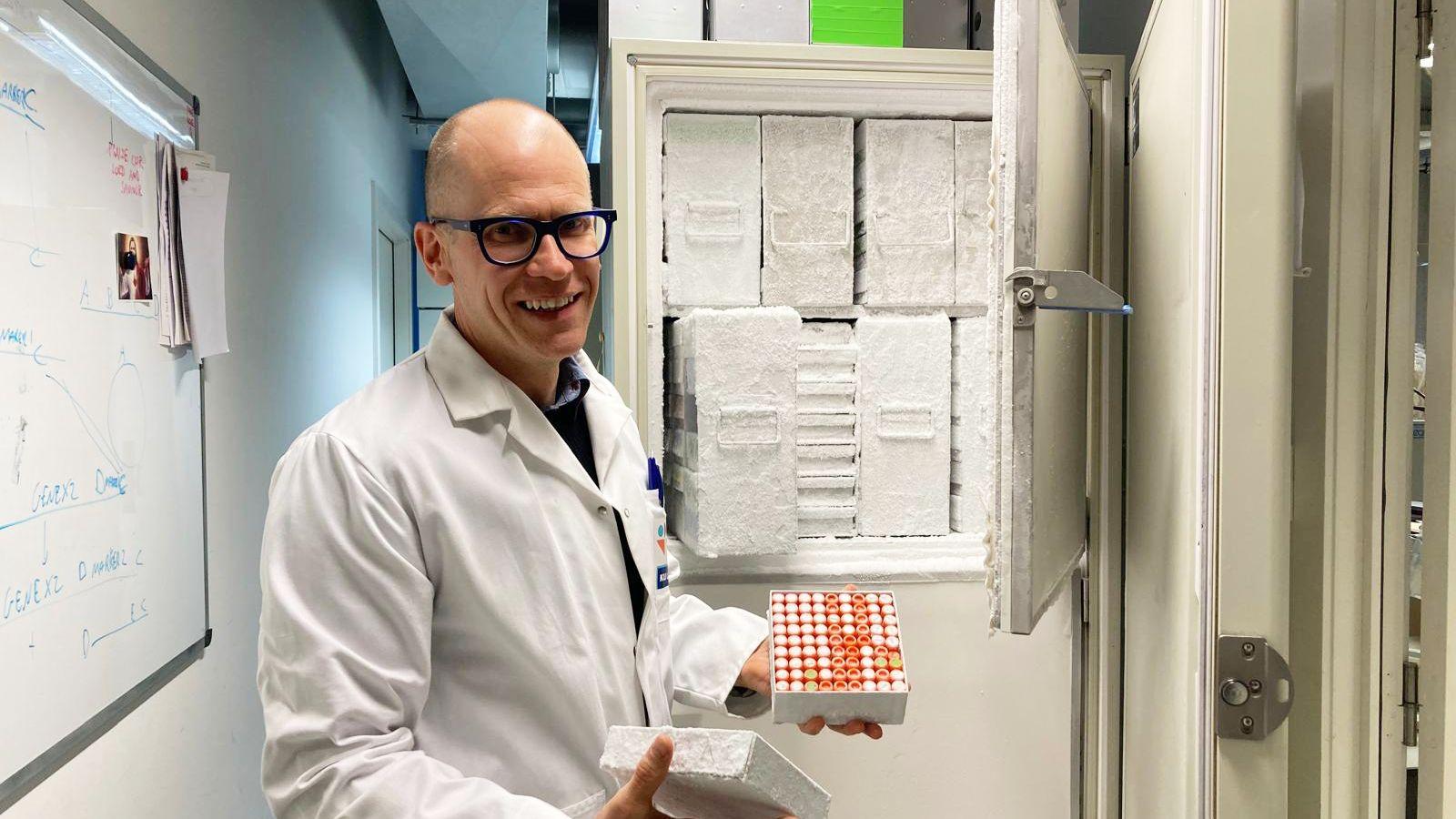 Čovek u belom laboratorijskom mantilu i crnim naočarima osmehuje se u kameru dok stoji ispred visokog otvorenog zamrzivača punog kontejnera; drži metalnu kutiju prekrivenu mrazom koja sadrži mnogo malih bočica