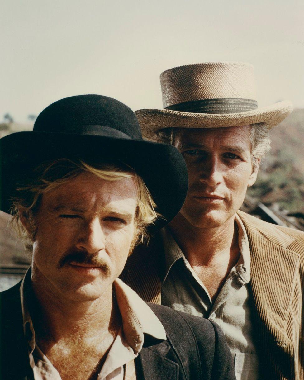 Robert Redford con bigote y un sombrero negro, detrás está Paul Newman con un sombrero beige