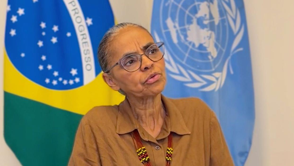 Marina Silva usando uma camisa marrom com as bandeiras do Brasil e da ONU atrás dela