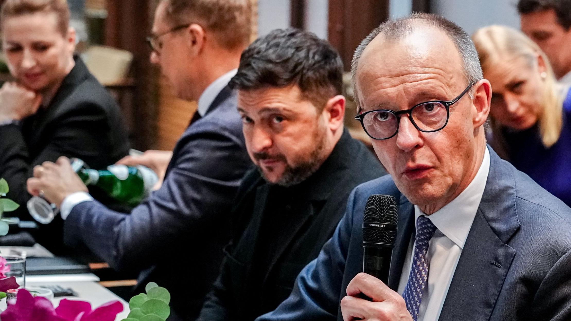 El canciller alemán, Friedrich Merz, habla mientras sostiene un micrófono, y está sentado a la mesa junto al presidente de Ucrania, Volodymyr Zelensky.