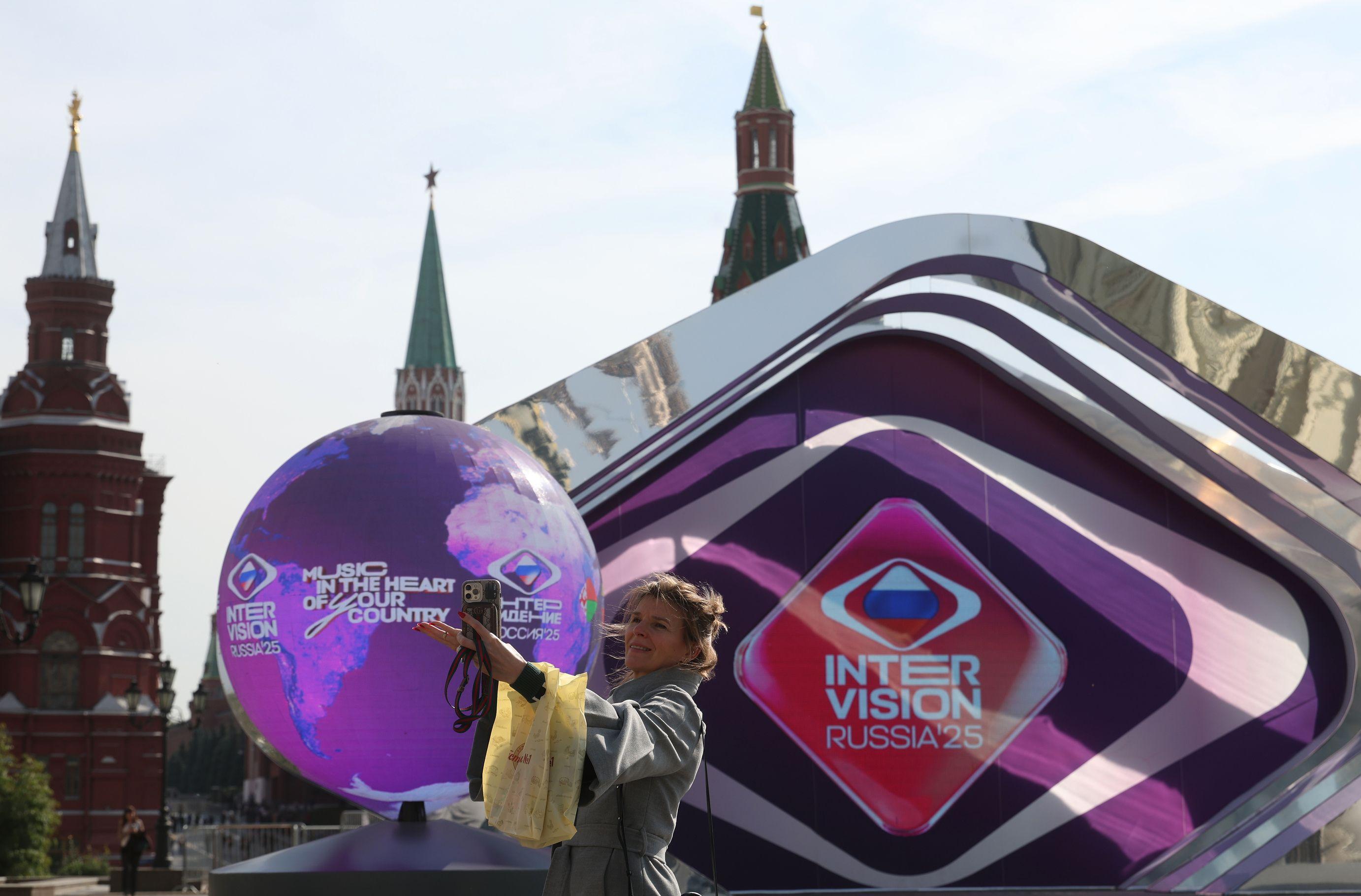 Una mujer se hace un selfi en la Plaza Roja de Moscú. Detrás de ella hay un gran globo decorativo y una pantalla con las palabras INTERVISION RUSSIA'25 y el eslogan "Música en el corazón de tu país".