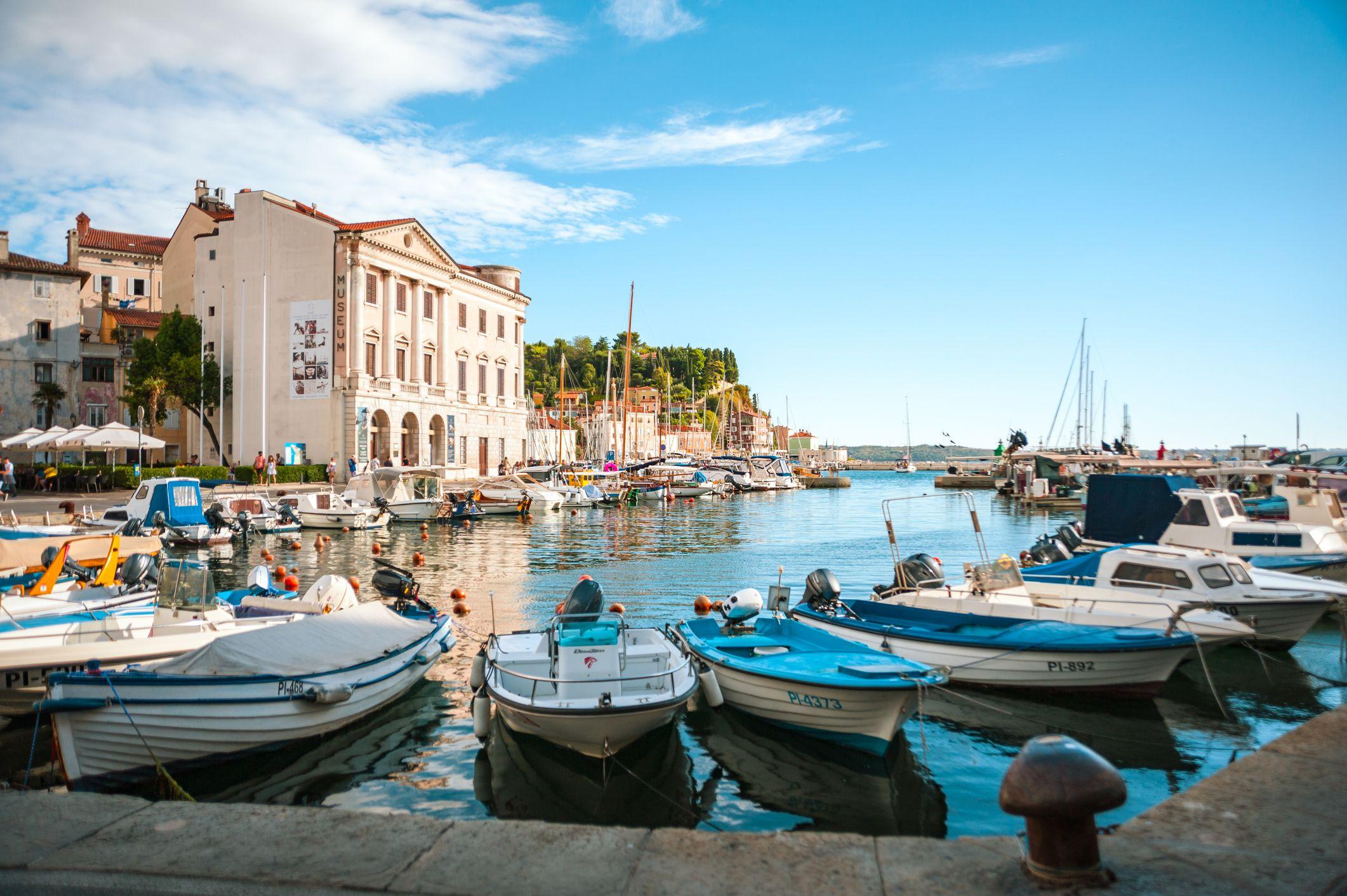 Piran, na Eslov&ecirc;nia. Porto com barcos, em meio &agrave; cidade hist&oacute;rica com seus edif&iacute;cios tradicionais, sob um c&eacute;u azul com algumas nuvens 