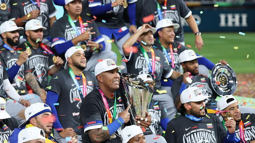 El equipo venezolano de béisbol todos con camisetas negras, gorras blancas, sentados juntos en mitad del campo de juego, con sus medallas y los trofeos. Del cielo caen papelillos de colores de celebración. 