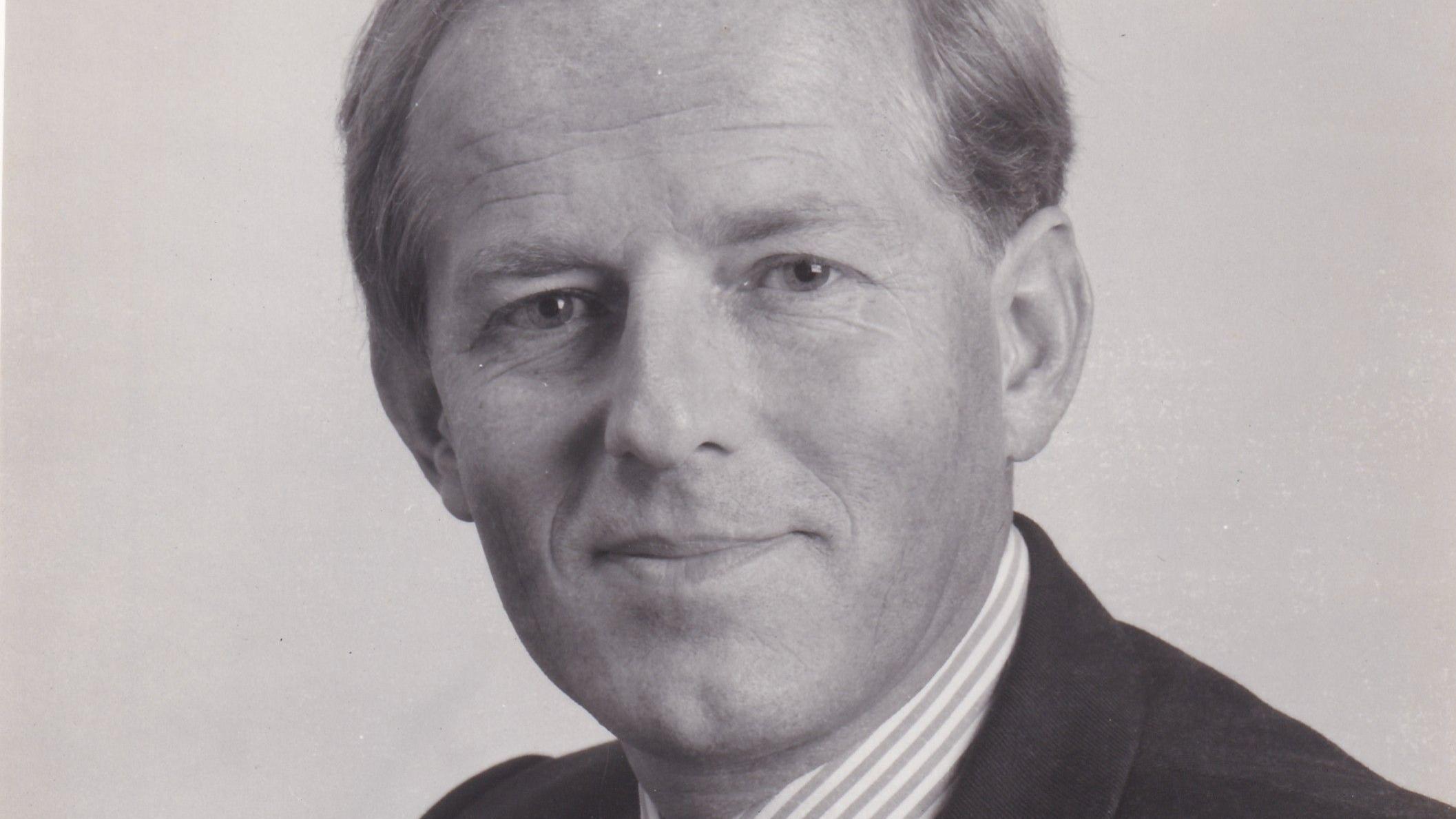 Retrato profesional en blanco y negro de John Smyth, quien viste una camisa de cuello a rayas y un traje oscuro. Sonríe a la cámara y parece amigable.