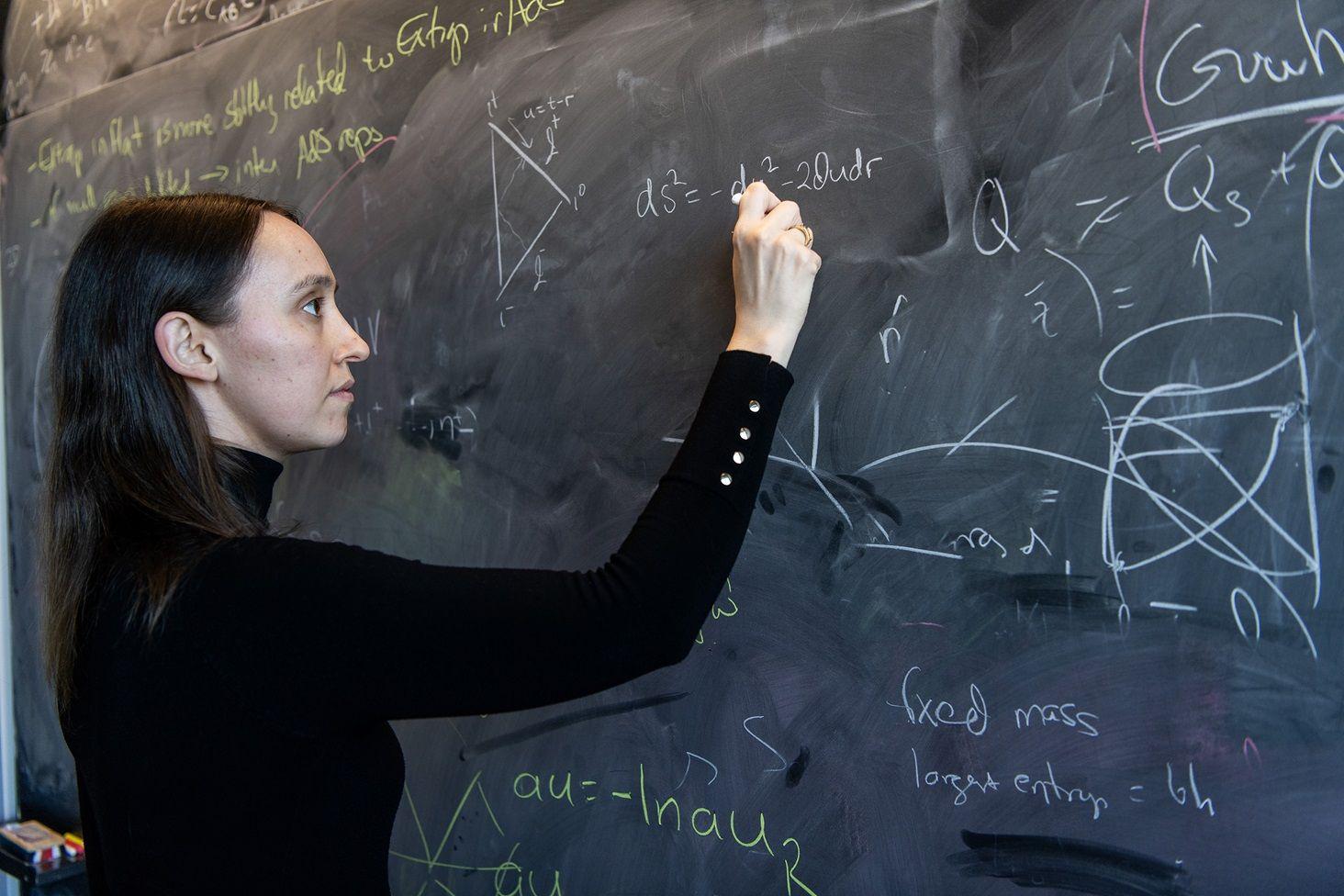 Sabrina González Pasterski con un saco negro escribe sobre un pizarrón lleno de fórmulas