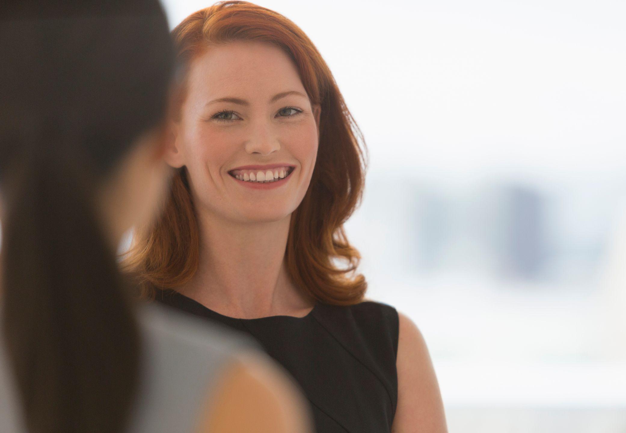 سيدة أعمال مبتسمة ذات شعر أحمر تتحدث مع زميلتها- صورة فوتوغرافية