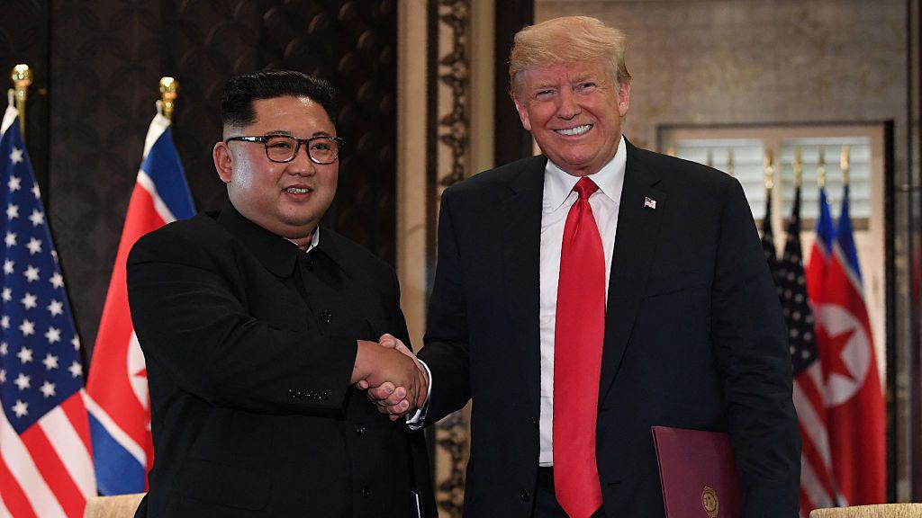 &Agrave; esquerda, Kim Jong-un, em um terno escuro estilo &ldquo;Mao&rdquo;; &agrave; direita, Trump, de terno preto e gravata vermelha. Os dois l&iacute;deres sorriem e apertam as m&atilde;os diante das bandeiras dos EUA e da Coreia do Norte