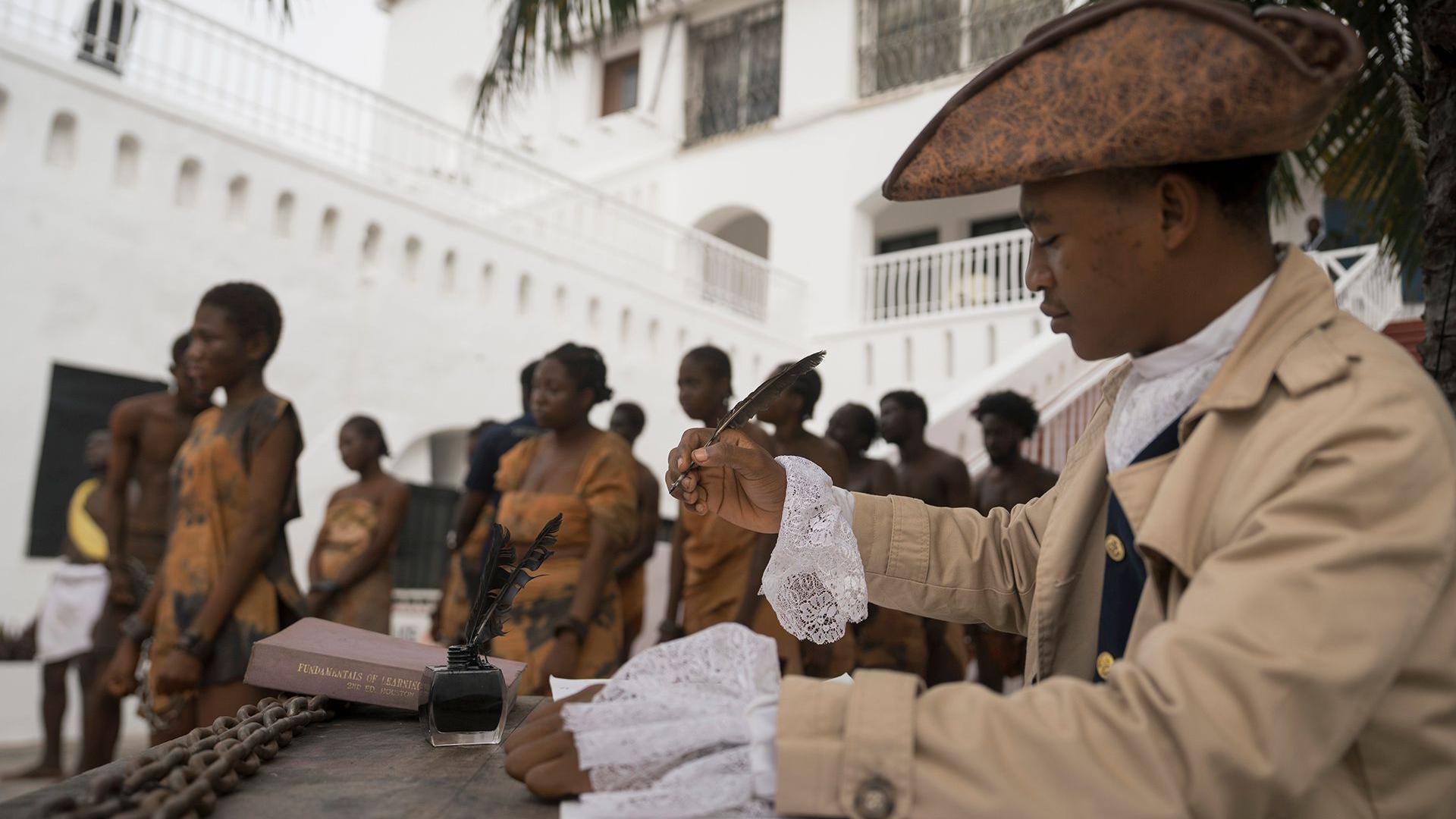 En primer plano, una persona viste ropa de la época previctoriana con sombrero y firma un documento con una pluma mientras que al fondo, personas vestidas con escasas túnicas de esclavos caminan encadenadas por la calle, durante una recreación del comercio transatlántico de esclavos en el patio del Castillo de Christiansborg en Acra, Ghana, el 19 de junio.