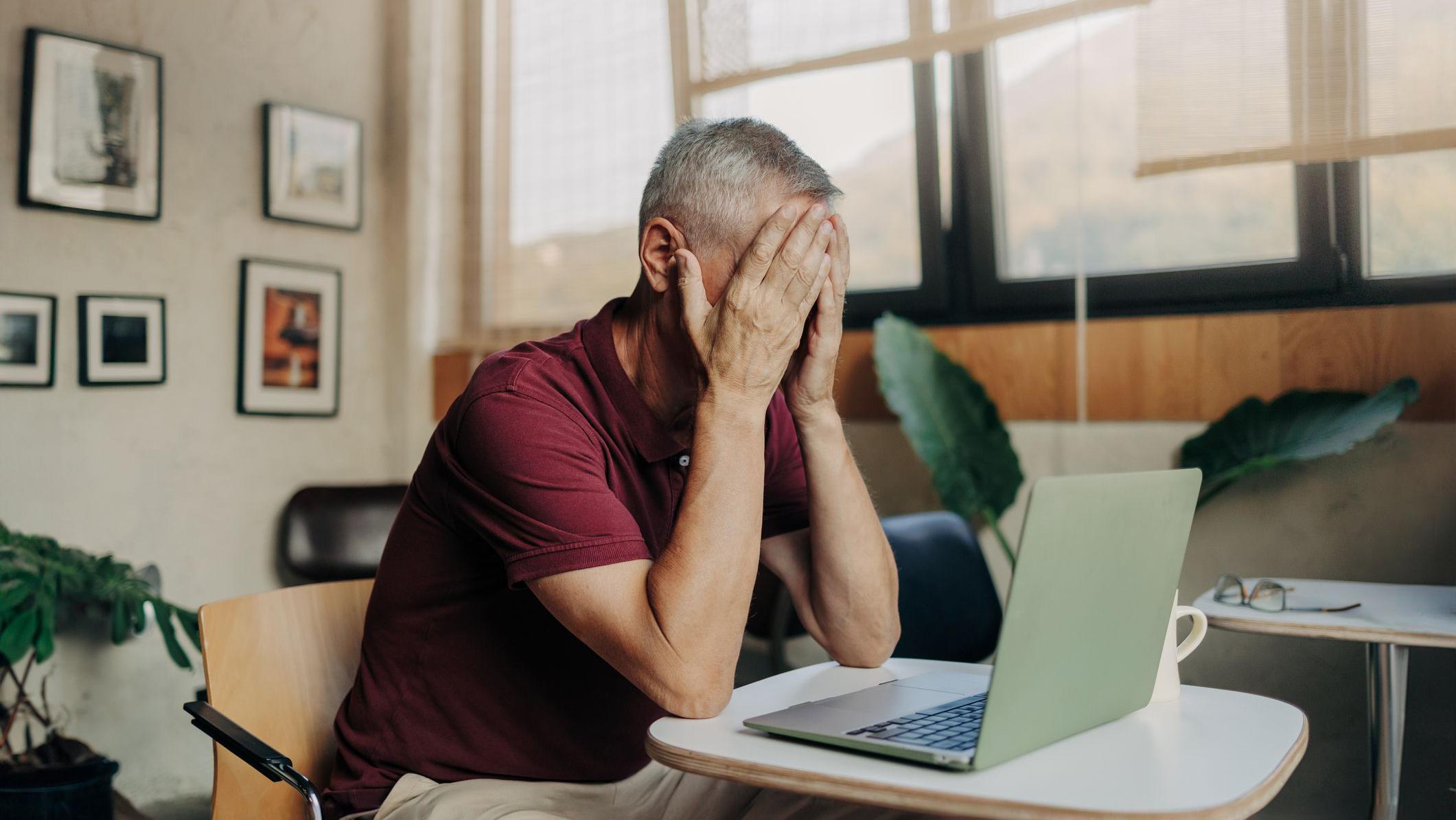 un hombre canoso, sentado en un escritorio y con un computador en frente, se lleva las manos a la cara. Aunque no se le ve el rostro su posición implica que está angustiado.