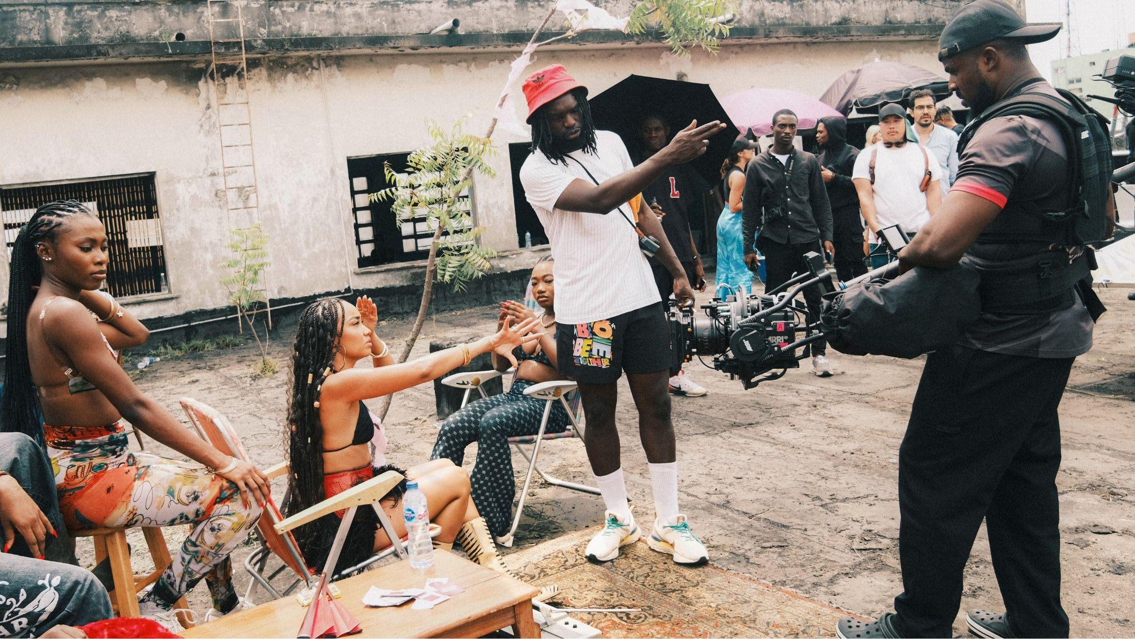 Meji Alabi, con un sombrero rojo, una camiseta blanca, pantalones cortos negros y calcetines y zapatillas blancas, sostiene una cámara y señala durante una sesión fotográfica. A su lado hay mujeres sentadas en tumbonas junto a un árbol delgado. Un camarógrafo, con una gorra negra y vestido con ropa oscura, está de pie al otro lado y el personal de producción, algunos con sombrillas negras, está detrás.