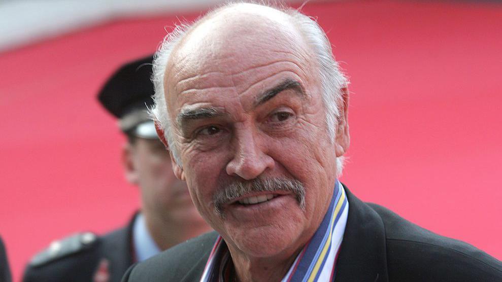 Sean Connery, de bigode, vestindo terno preto e camisa listrada