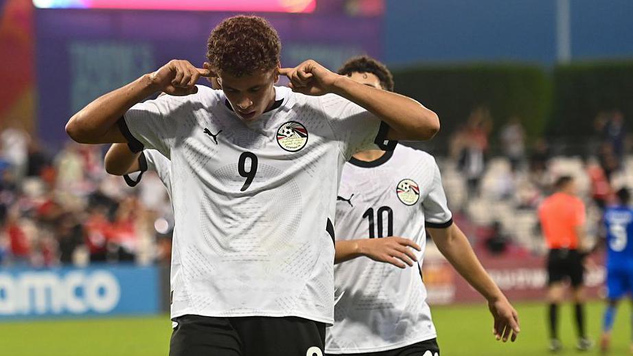 حمزة عبد الكريم مهاجم منتخب مصر يحتفل بتسجيل هدف
قميص أبيض رقم 9، وسروال أسود، يضع أصابعه في أذنيه، وخلفه يظهر قميص لاعب رقم 10