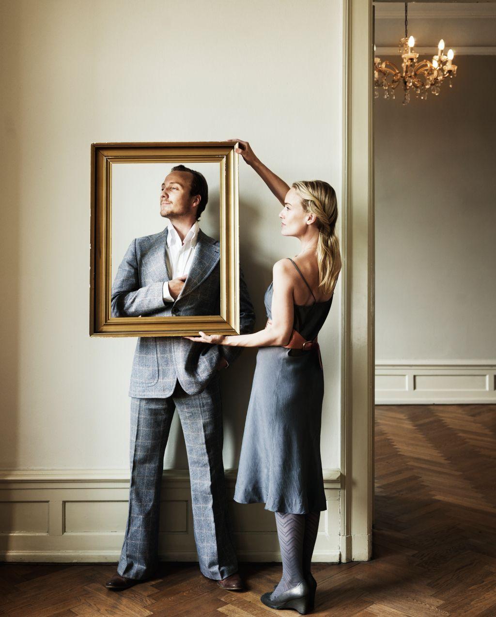 Mujer vestida de gris le pone un marco dorado a un hombre que está recostado a una pared, posando, en un apartamento elegante. 