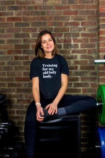 Elizabeth Davies sits on a fitness block in a gym, which has exposed brick and weights that can been seen at the edges of the photo. She crosses one leg on top of the block while the other hangs off. She is smiling and wears dark leggings and shoes, and a t-shirt that reads: 'Training for my old lady body.' She has shoulder length brown hair.