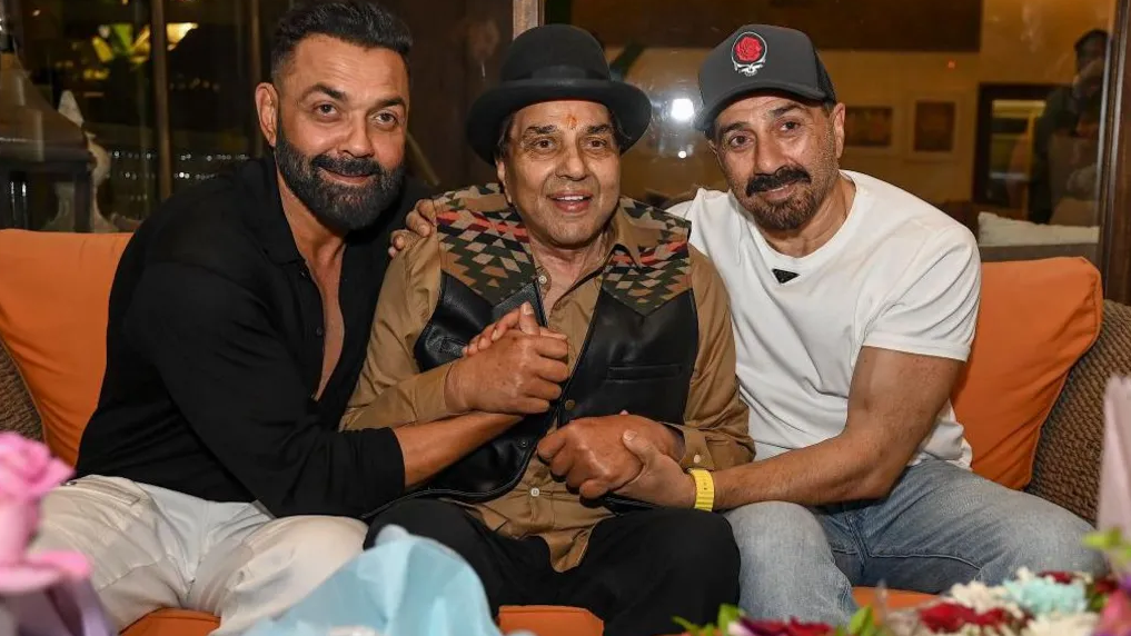 النجم دارمندرا (في الوسط) مع ولديه ساني (يمين) وبوبي ديول (يسار) في مقر إقامته في مومباي. دارمندرا بعد أن كبر في السن ويرتدي قبعة سوداء وقميصاً عسلي اللون وجاكيت بدون أكمام أسود وبنطالاً أسود في المنتصف بين ابنيه الشابين أحدهما (ساني) يرتدي قميصاً أسود والآخر قميصاً قصير الأكمام بلون أبيض.