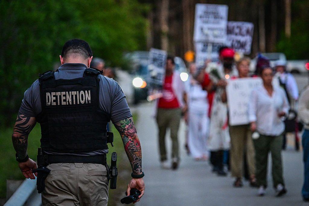 Un funcionario de prisiones pasa de espaldas junto a personas que sostienen velas, pancartas y flores durante una vigilia afuera del Centro de Detención Krome en Miami el 24 de mayo de 2025, protestando contra la custodia de Inmigración y Control de Aduanas de EE. UU. y las deportaciones masivas. (Foto de GIORGIO VIERA/AFP a través de Getty Images)