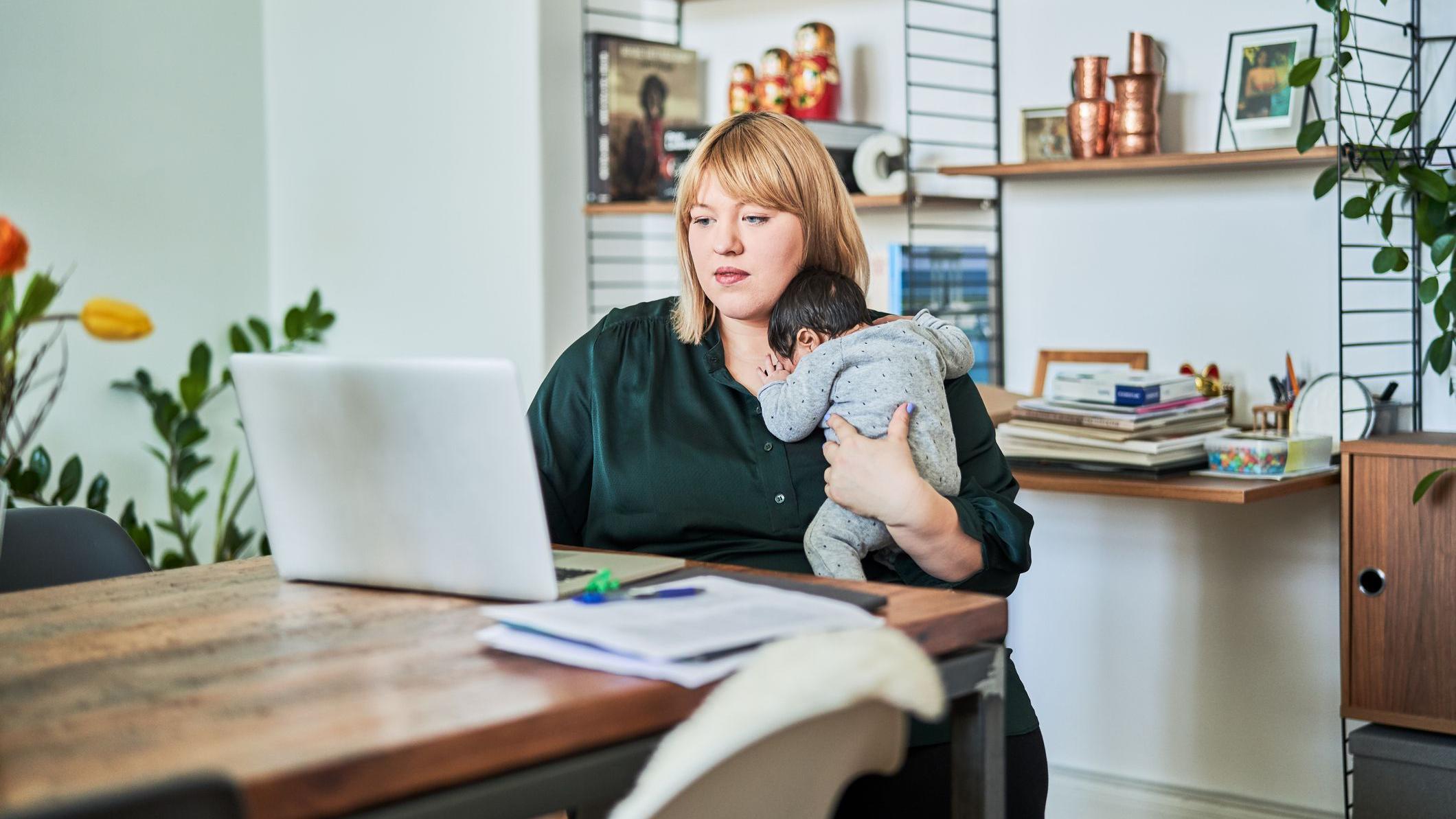 Foto de uma mulher sentada em uma mesa em frente a um computador com um bebê no colo. 