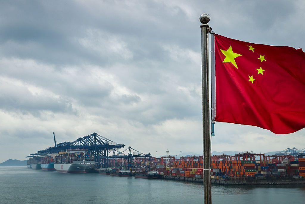 La bandera china ondea en un puerto y al fondo se vende contenedores de carga.