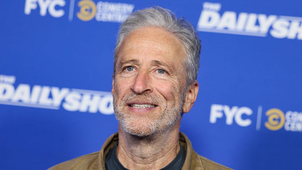 Jon Stewart, de jaqueta marrom e camiseta preta, com fundo de cor azul promocional do seu programa 'The Daily Show'