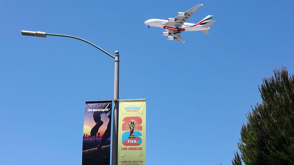 Bandeiras da Copa do Mundo de 2026 em um poste de Los Angeles, nos Estados Unidos, com um avi&atilde;o de passageiros voando no c&eacute;u azul 