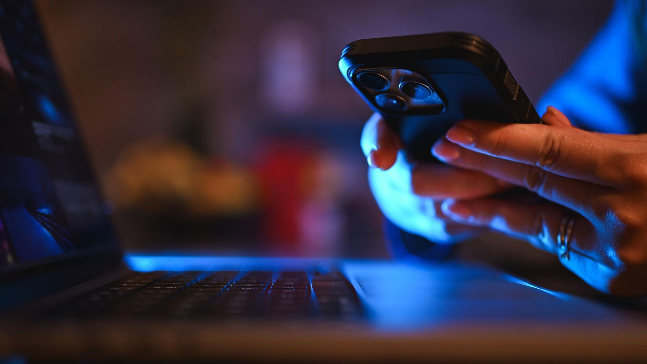 M&atilde;os seguram um telefone celular em frente a um laptop, sob luz azul