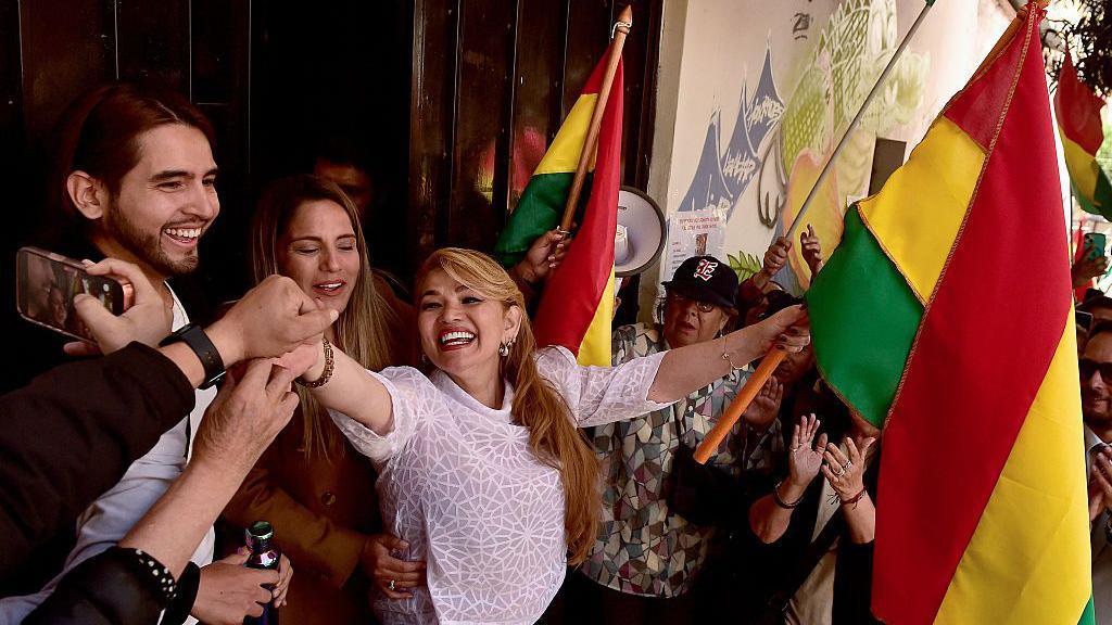 Jeanine Áñez sonriente mientras sostiene una bandera de Bolivia con una mano y saluda a la gente con la otra.