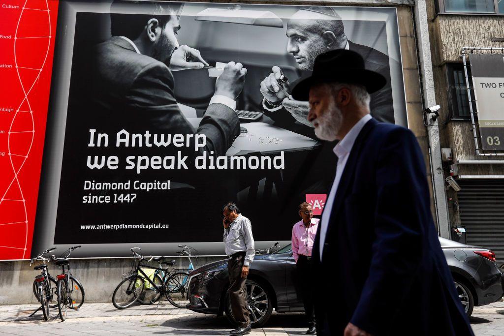 Escena urbana en Amberes con varias personas caminando frente a un gran cartel publicitario que dice: ‘In Antwerp we speak diamond – Diamond Capital since 1447’ ("En Amberes hablamos el idioma de los diamantes: Capital mundial del diamante desde 1447"). Muestra una imagen en blanco y negro de dos personas examinando diamantes. Hay bicicletas estacionadas junto a la acera y un automóvil estacionado frente al cartel.