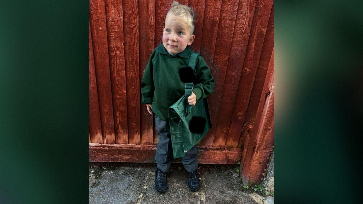Edward en uniforme escolar verde, con un maletín y está parado frente a una valla de madera  