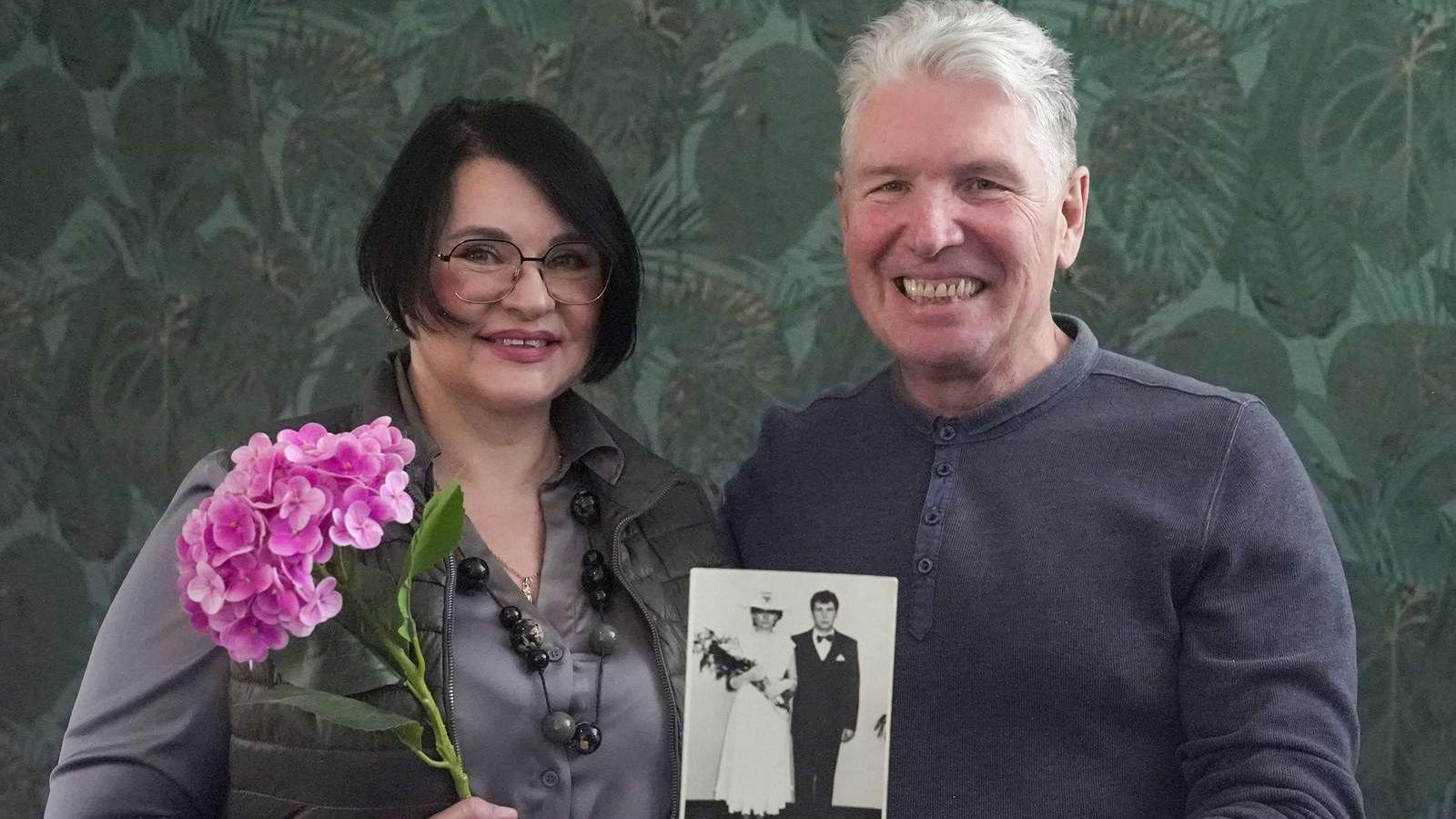 Muškarac i žena stoje jedan pored drugog. Ona drži ružu u ruci a on fotografiju