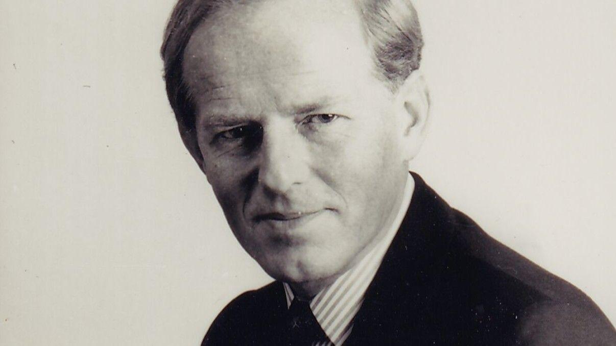 Una fotografía en blanco y negro de John Smyth con una camisa a rayas y un traje oscuro.