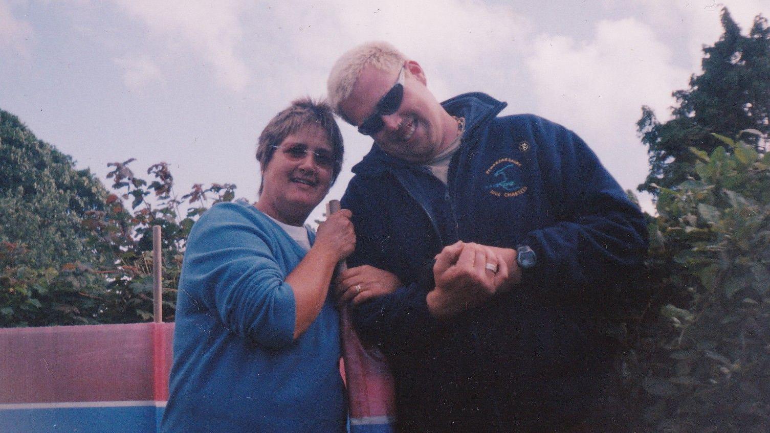 Photograph of Dale Morgan (right) and his mother Judith Rhead (left). They stand arm in arm, Judith holds a windbreaker pole with her left hand between them, and links her right arm to Dale. She wears tainted thin sunglasses and a blue v neck jumper. Dale wears runner black tinted sunglasses and a navy long sleeve zip up fleece. Blue skies can be seen behind them. 