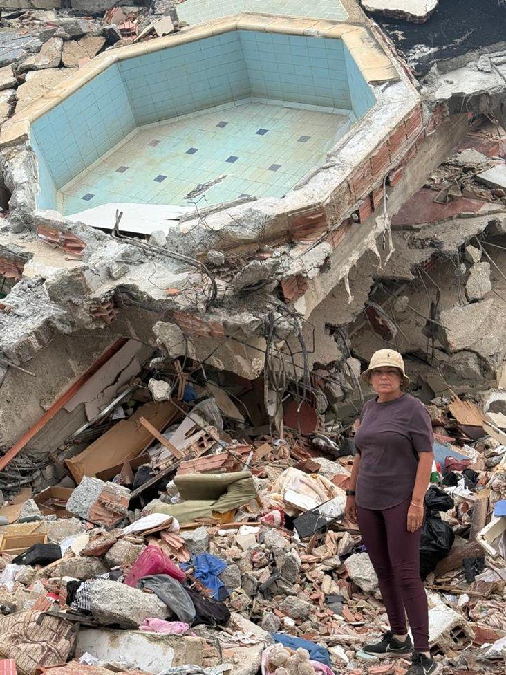 María Lourdes Pérez, vestida con un pantalón y camisa marrones y un gorro beige, está parada en una zona llena de escombros. Detrás de ella está lo que parece ser una piscina pequeña, piedras, hormigón, partes de una edificación, muebles destruidos, ropa.

