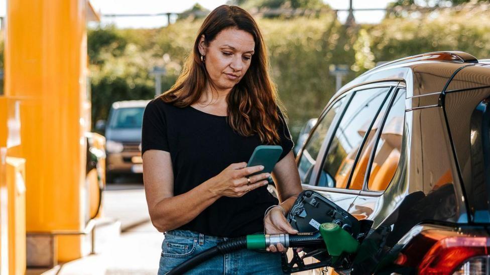 Una mujer llenando el tanque de su automóvil en una estación de servicio mientras consulta su teléfono móvil