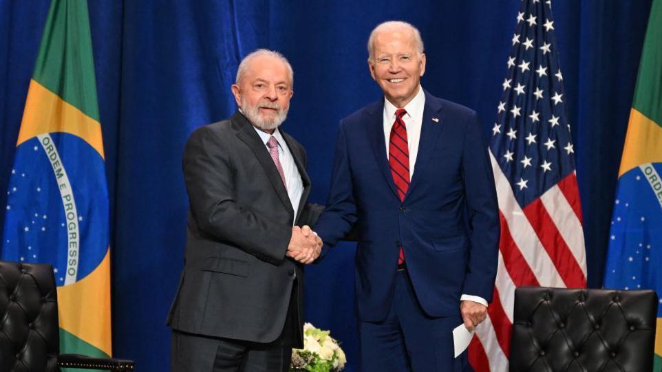 Lula y Joe Biden se dan un apretón de mano mientras miran a la cámara. Ambos visten trajes oscuros. En el extremo izquierdo se ve la bandera de Brasil y en el derecho, la de Estados Unidos.