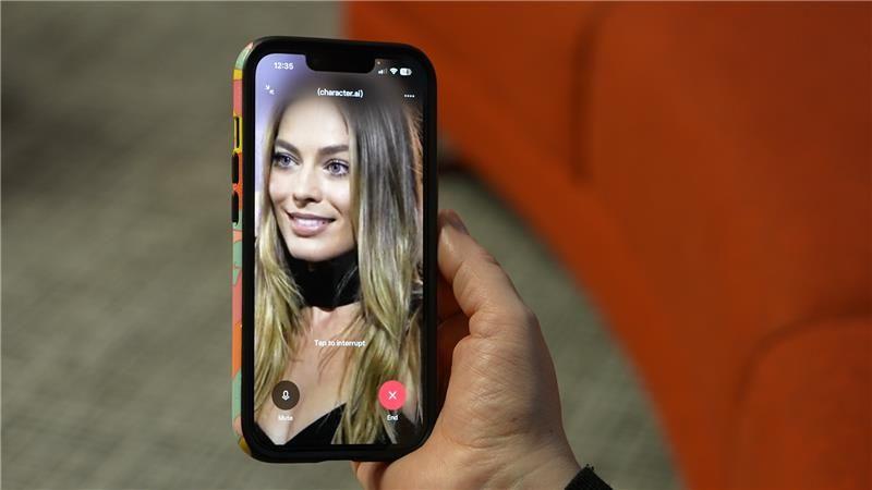 A still image of the actress Margot Robbie on a phone screen which is being held in from on an orange sofa. 