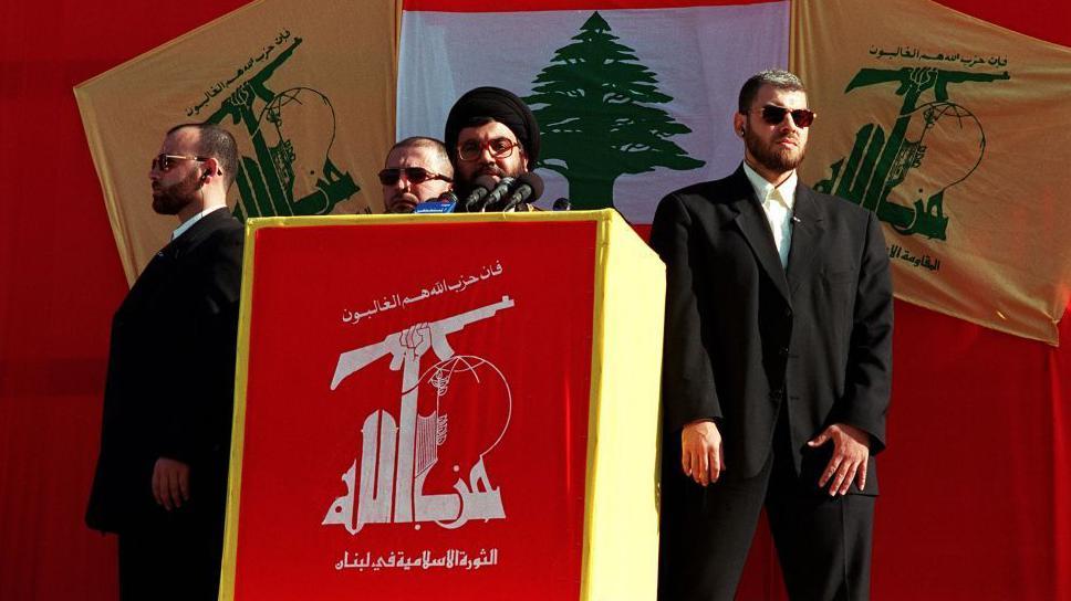 أمين عام حزب الله السابق حسن نصرالله يلقي كلمة محاطا بثلاث رجال أمن باللبتاس الأسود مع خلفية باللون الأحمر وعلم لبنان وعلم حزب الله
