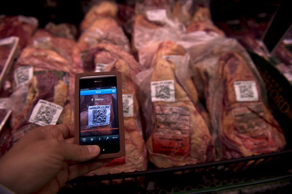 Una bandeja con cortes de carne vacuna envasada que tienen un código QR para obtener su trazabilidad, y delante una persona sostiene con su mano un teléfono y apunta con la cámara a uno de esos códigos.
