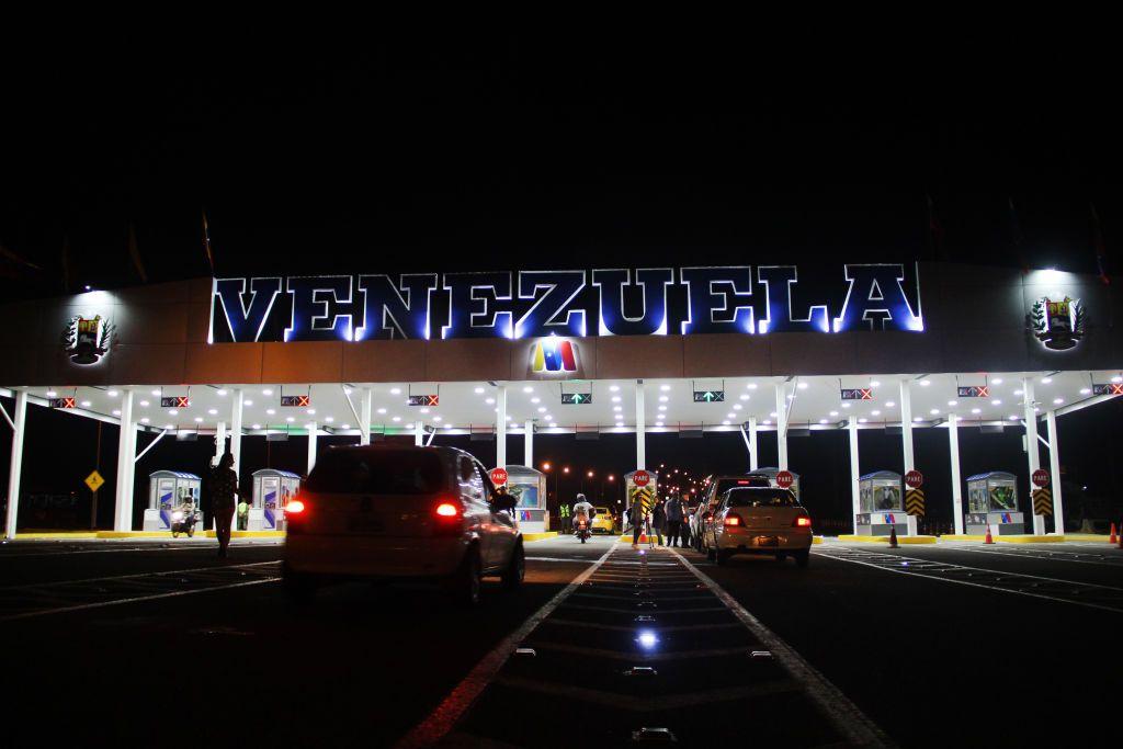 Peaje fronterizo iluminado de noche con el letrero “Venezuela”, mientras varios autos avanzan hacia los puestos de control.