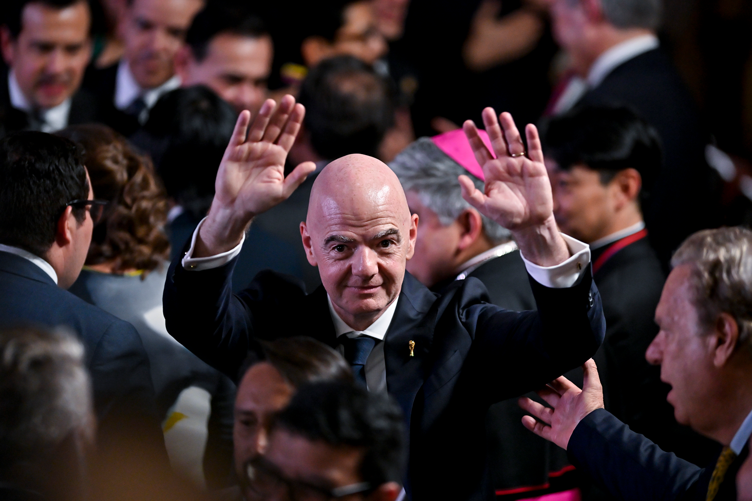 Gianni Infantino, vestido con una camisa blanca y una corbata y traje oscuro, sube los brazos en medio de varias personas

