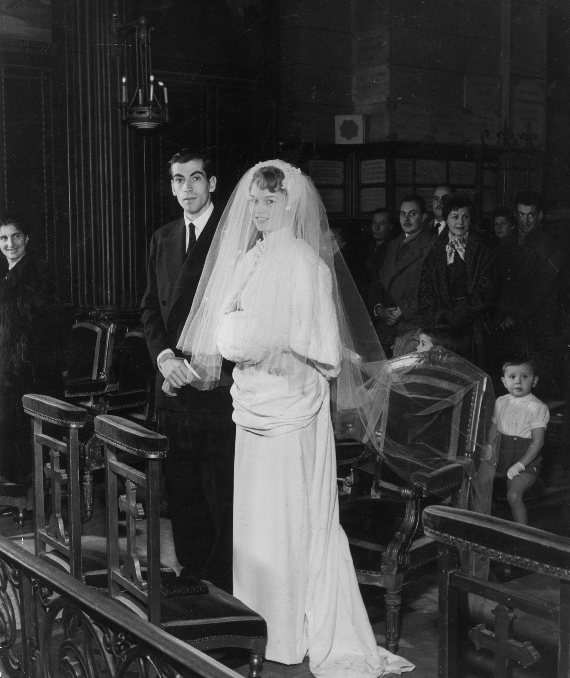 Brigitte Bardot e Roger Vadim, em p&eacute; diante do altar durante a cerim&ocirc;nia de casamento em dezembro de 1952. O rosto de Bardot est&aacute; coberto por um longo v&eacute;u e Vadim usa um terno escuro e gravata.