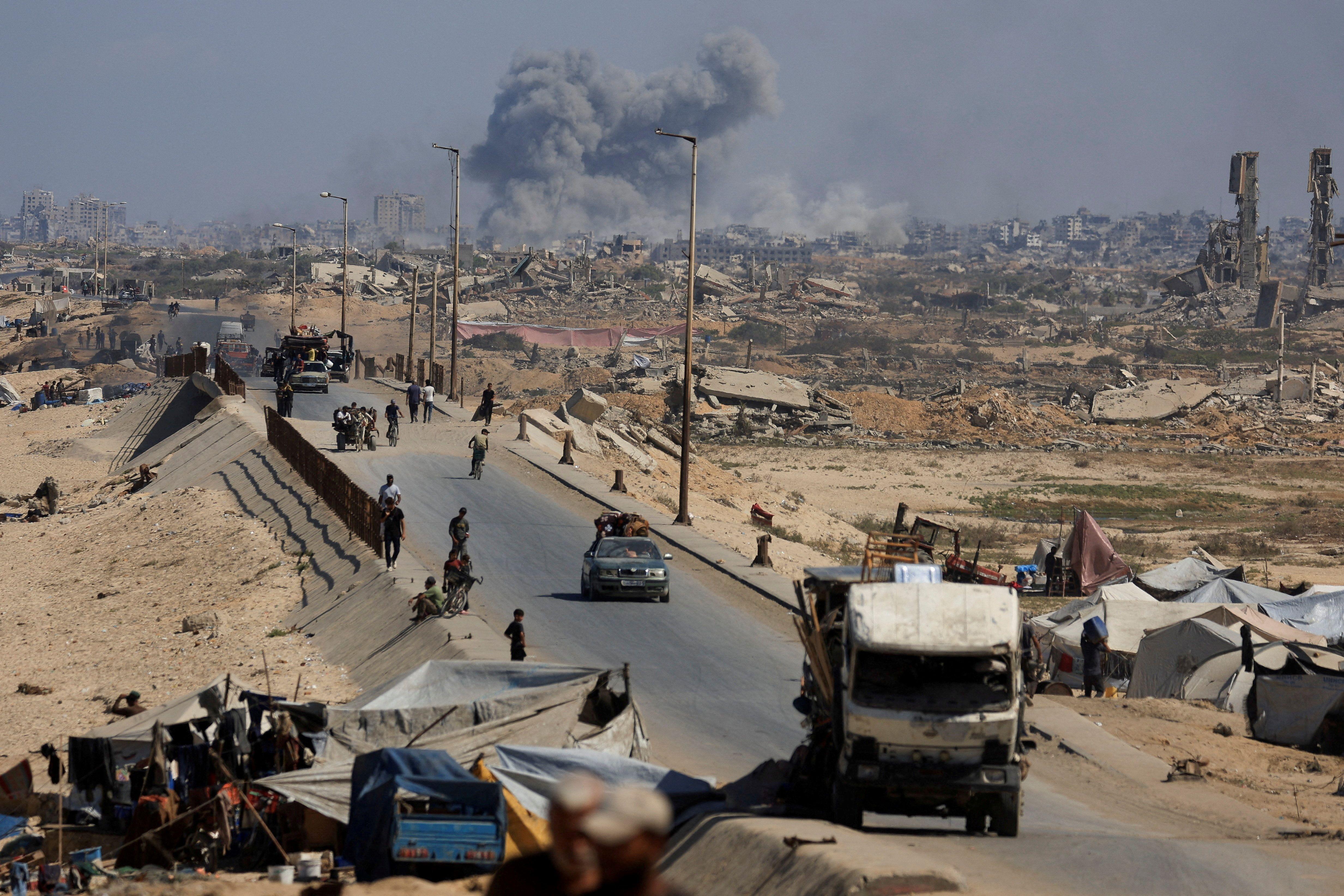 Una carretera por la que pasan palestinos desplazados y al fondo la Ciudad de Gaza, destruida, de la que asciende una gran nube de polvo de un bombardeo israelí.