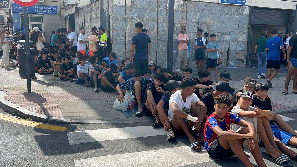 Un grupo de niños y jóvenes hacen fila sentados en mitad de una calle de Ceuta. 