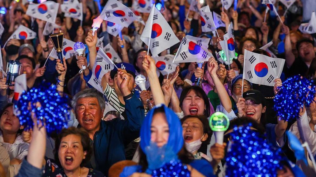 2025年6月3日，在南韓首爾，民主黨總統候選人李在明的支持者手持K-Pop應援棒與韓國國旗，聚集在國會外，當時正值開票進行中
