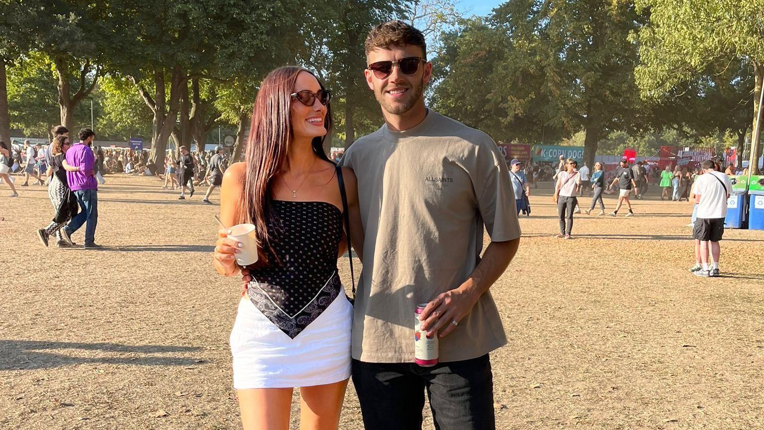 Lauren Macpherson aparece ao lado do namorado, Zak. Os dois est&atilde;o em um festival. Ela tem cabelos longos e escuros e usa uma saia branca e um top preto tipo bandana, enquanto ele veste uma camiseta bege e jeans pretos