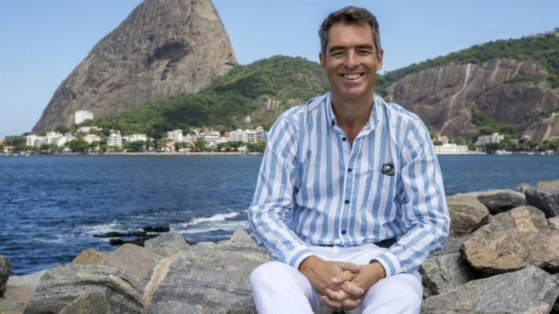 Tim Vickery sentado em rochas com o o mar e os morros do Rio de Janeiro ao fundo