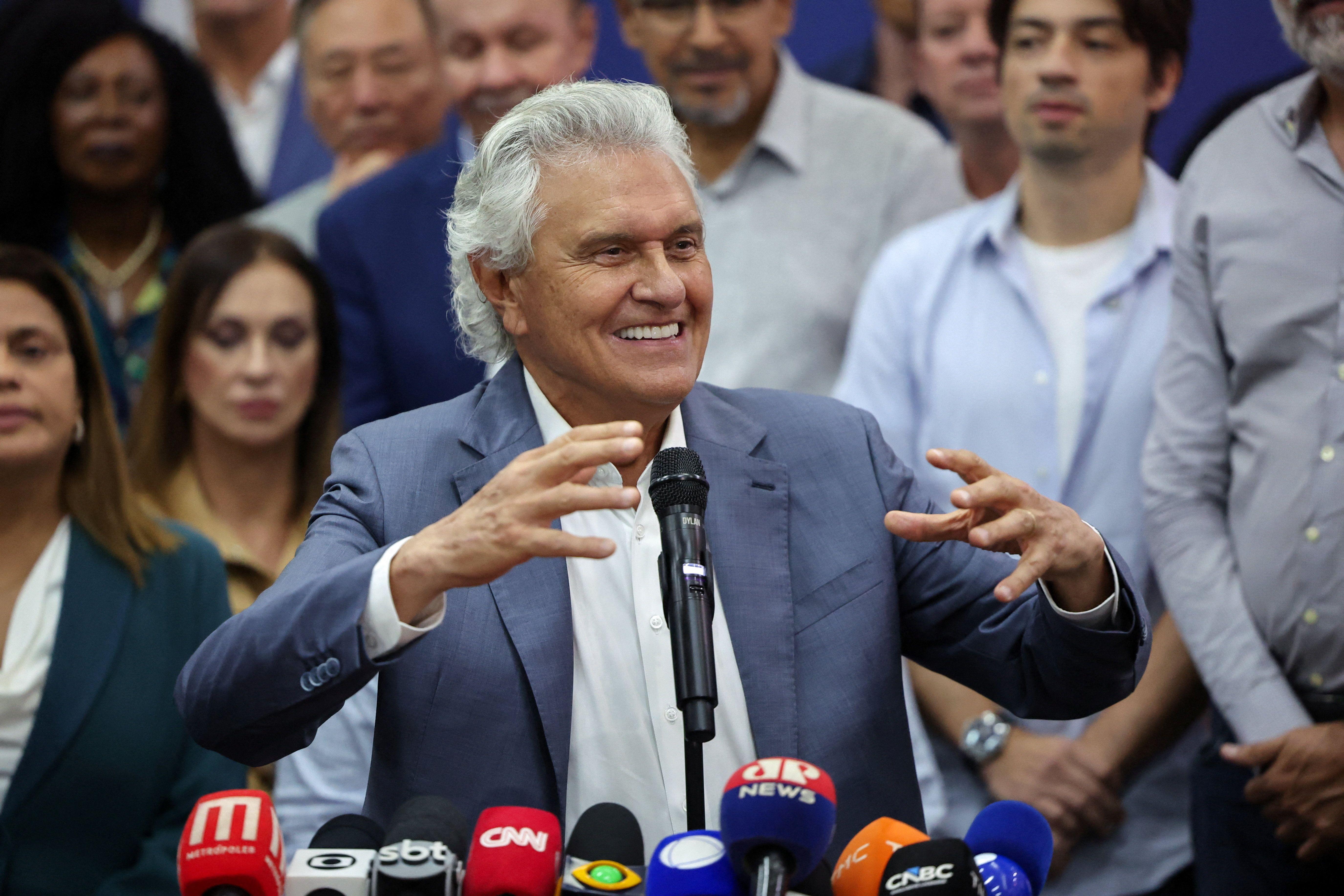 Caiado de blazer azul gestocula ao discursar ao microfone para apoiadores cercado de aliados pol&iacute;ticos