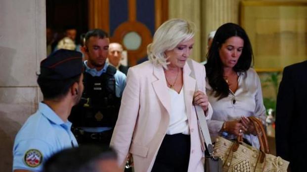 Marine Le Pen, una mujer rubia con chaqueta rosada, camisa blanca y falda negra, sale del Tribunal de Apelaciones en Francia, acompañada de una asistente y seguida de un guardia con chaleco antibalas.