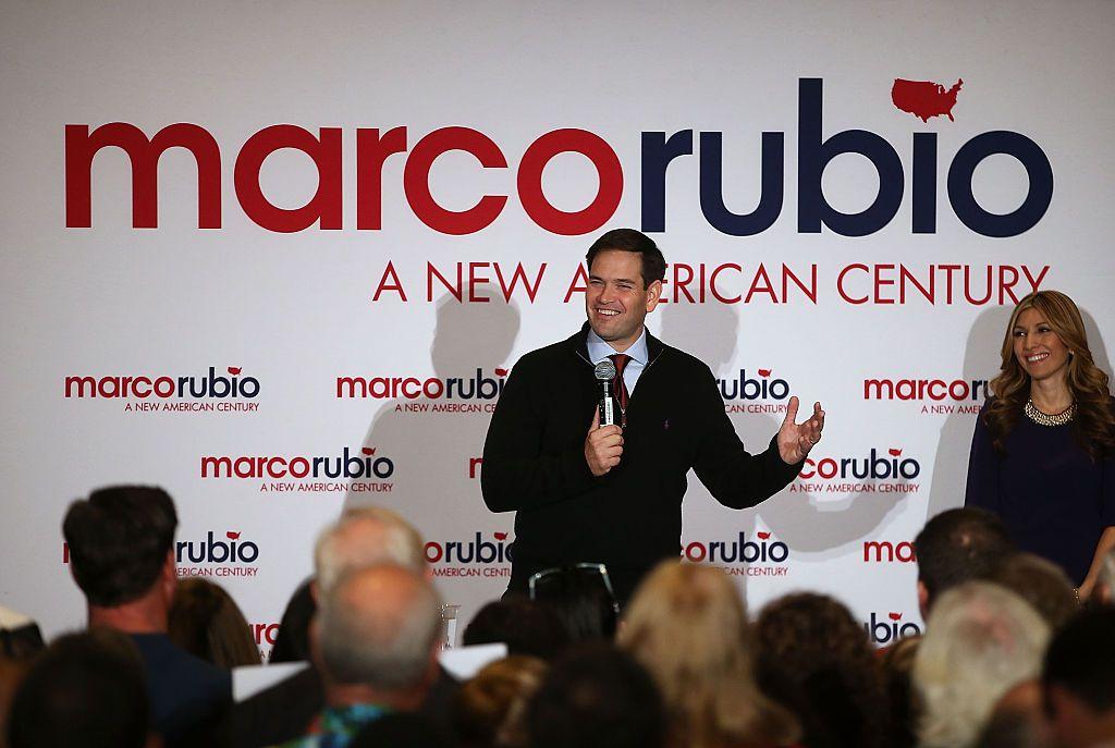 Marco Rubio falando no microfone. Ao fundo, há um painel com o nome dele