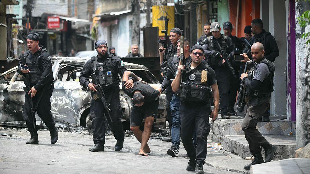 Grupo de policías con un sospechoso, en foto en la calle. Están fuertemente armados y uniformados. El sospechoso lleva gorra, mira al suelo y está descalzo.
