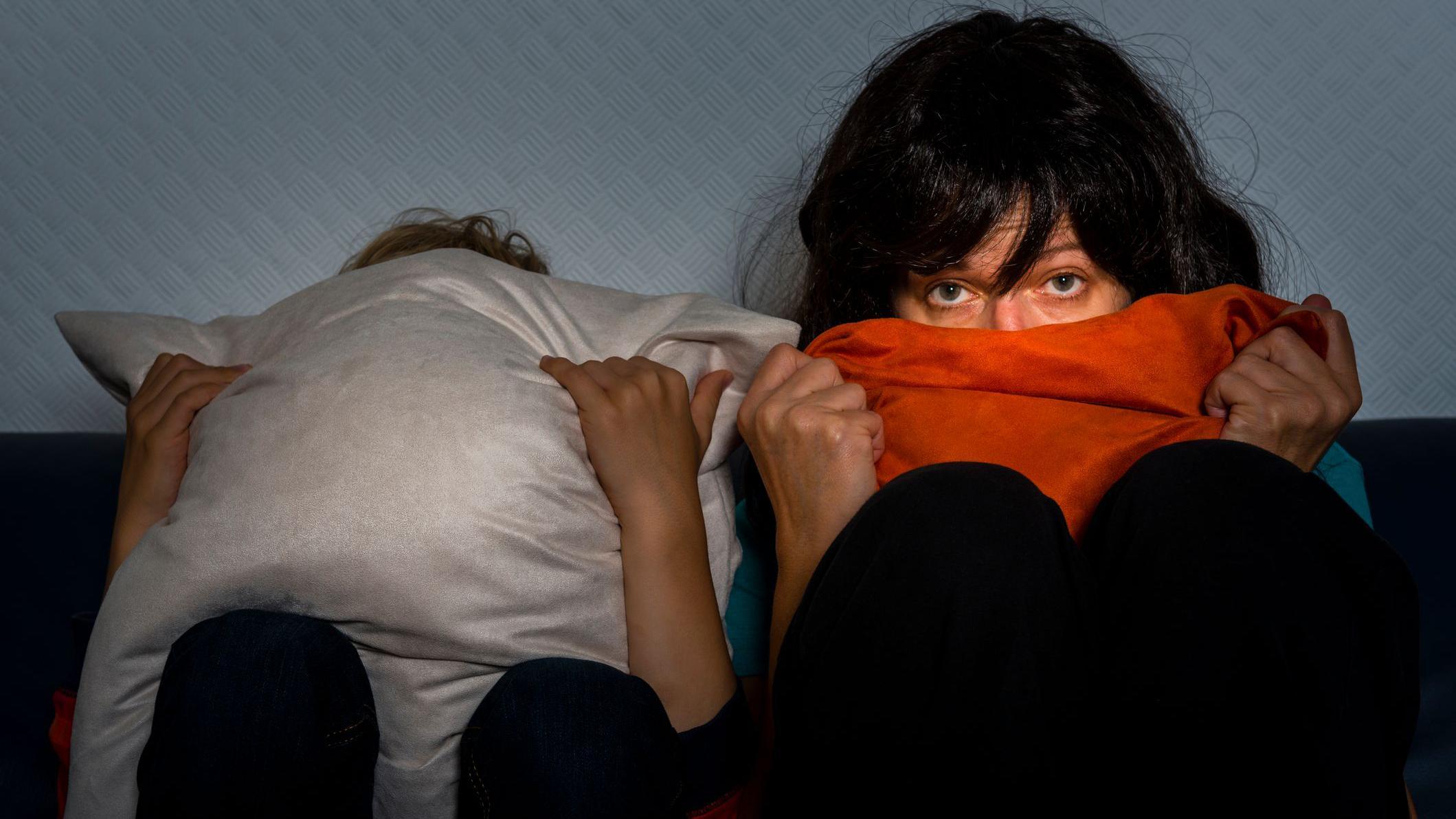 Duas mulheres vendo televisão sentadas na cama. Uma delas se cobre totalmente com uma almofada e a outra leva o cobertor à boca, mas continua assistindo atentamente com os olhos assustados